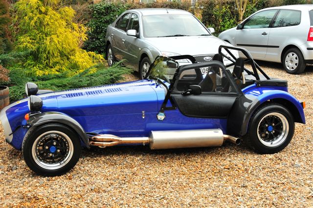 Any Seven owners in Barcelona? - Page 1 - Caterham - PistonHeads - This image depicts a colorful, vintage-style vehicle parked on a gravel lot. The vehicle is painted in a combination of blue and black, with a convertible top that appears closed. It features a prominent front grille and is equipped with black wheels and wheel wells. In the background, there are other vehicles parked, including one silvertone car and a second vehicle that is partially obscured. The scene suggests a relaxed outdoor setting, possibly a parking area or a lot for shows or events.