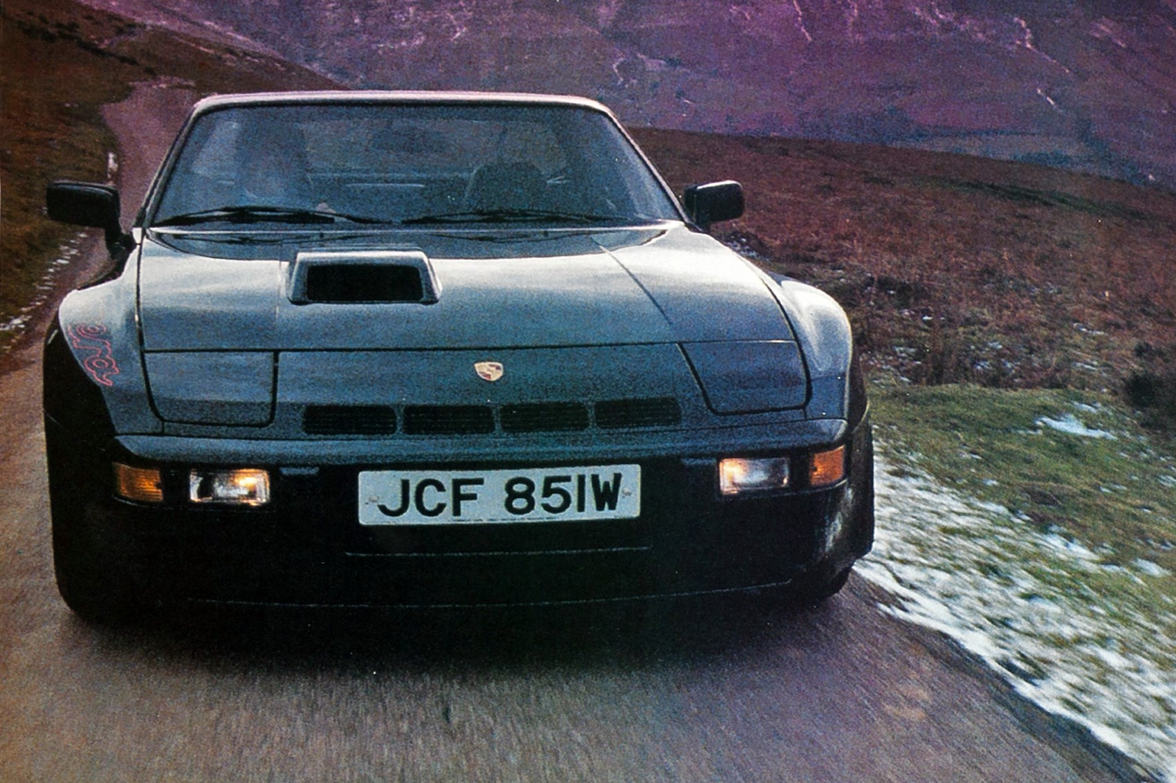 So what car have you been obsessing about today?? (Vol 2) - Page 422 - General Gassing - PistonHeads - This image captures a modern, black sports car driving on a winding road. The car is a Porsche, distinguishable by the unique emblem on the grille. It's positioned slightly off-center, moving from the right side to the left. The setting appears to be a mountainous area covered in a thin layer of snow on the side of the road.