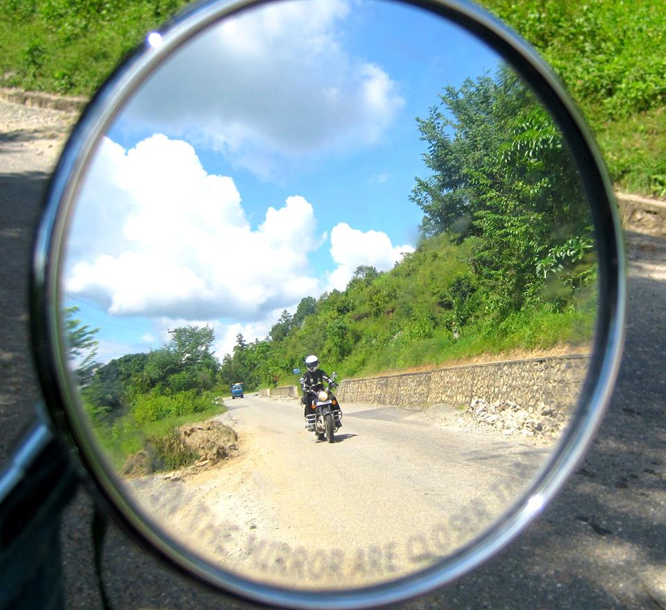 Nepal, Bhutan, India - Page 3 - Biker Banter - PistonHeads - The image shows the reflection of a motorcyclist in a round rearview mirror. The motorcyclist is seen riding on a paved road that is lined by lush green trees. The sky above is a clear blue with scattered white clouds, suggesting a pleasant day. The surrounding scenery suggests a landscape possibly in the countryside or on the edge of forested areas. The reflection in the mirror contains the motorcyclist, their motorcycle, and a portion of the road, capturing a moment of outdoor activity.