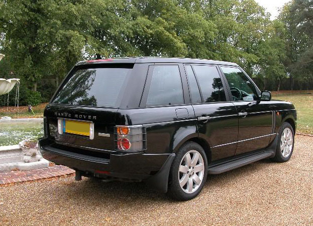 Sell Range Pistonheads Rover - The image depicts a sleek black Range Rover parked on a gravel surface. The car is oriented towards the right of the frame, showcasing its chrome grille, angular headlights, and distinctive tail lights. The vehicle's glossy finish reflects a water feature in the background, adding a serene touch to the rural setting. The scene is framed by trees and a hint of a fountain, suggesting an upscale residential area.