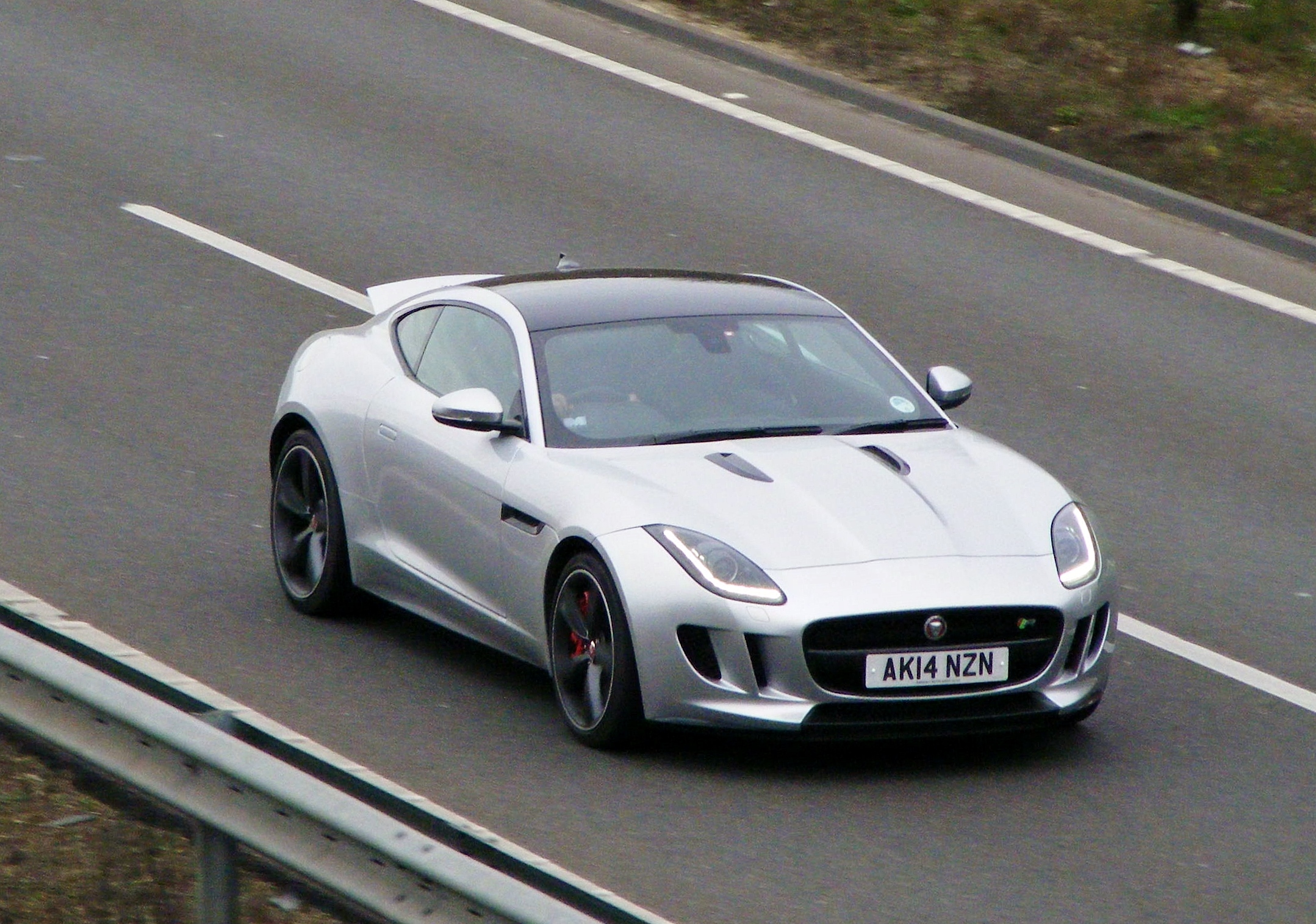 Herts, Beds, Bucks & Cambs Spotted - Page 366 - Herts, Beds, Bucks & Cambs - PistonHeads - The image shows a silver Jaguar sports car in motion on a two-lane road. The car is sleek and modern, with a low and aerodynamic profile. The license plate of the car reads "A14 NZN". In the background, there is a guardrail on the side of the road, indicating a margin of safety for motorists. The road appears to be a highway, as it's a high-speed road with markings for lanes and shoulders.