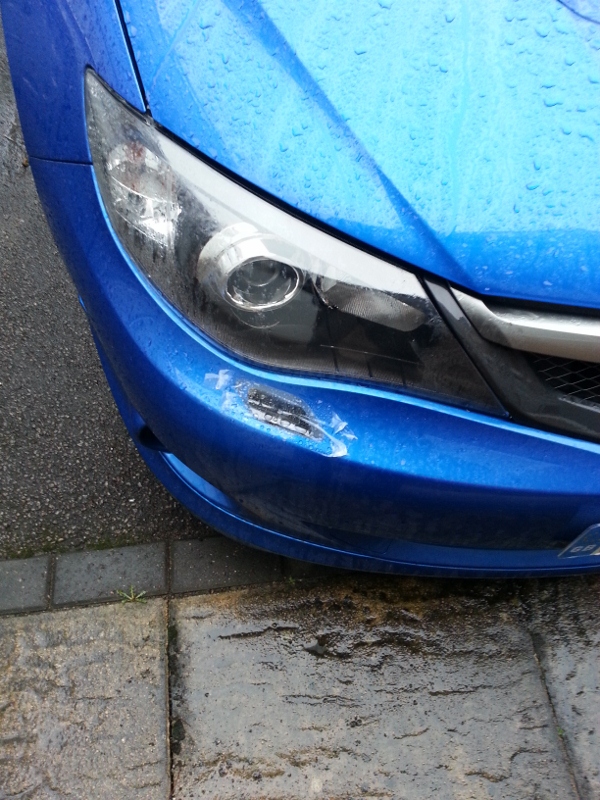 avoiding main dealer prices - Page 1 - Subaru - PistonHeads - The image shows a close-up view of a blue sedan, primarily focusing on its front bumper and hood area. The car appears wet, suggesting it has been raining. There is visible damage on the car's bumper, and the hood has a scratch mark. The headlight of the car is clean and reflective, indicating it has not been in contact with the pavement.