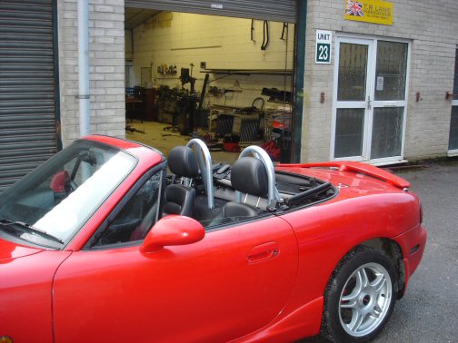 Throw away your style bar… - Page 1 - Mazda MX5/Eunos/Miata - PistonHeads - This is a photograph of a red sports car with its top down, parked in front of a garage. Inside the vehicle, one can see a pair of helmets resting on the passenger side's seats. The garage itself appears to be used as a workshop, with various equipment and parts visible through the garage door, and there are numbered items in the background. The street at the bottom is clear and no other vehicles are visible in the immediate area. This image suggests a scene of a garage where car activities do take place.