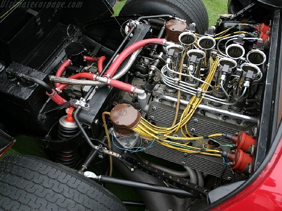 Fujima Alfa Romeo T33 stradale - picture heavy (possibly) - Page 1 - Scale Models - PistonHeads - The image shows a close-up view of an engine compartment with the hood of a car slightly open, revealing internal components. The engine appears to be a V8 with various hoses, wires, and mechanical parts visible. Red hoses that run across the motor add a contrasting color to the scene. The engine occupies the majority of the frame, indicating its significance in this photo. The overall structure of the engine block and the precise arrangement of its components, such as the connecting rods, support the identity of the V8.