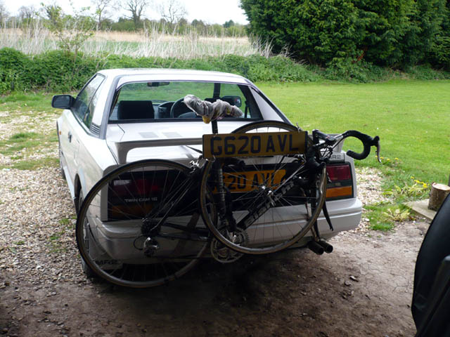 Cayman bike carrier ? - Page 1 - Boxster/Cayman - PistonHeads - In the image, a white car is parked on a patch of gravel, with a bicycle leaning against its trunk. The bicycle bears a yellow license plate that reads "G62 AVL", which suggests the car's registration or its corresponding tag. The car appears to be older, with a noticeable left rear wheel well. Alongside the car are lush green bushes, creating a natural backdrop. The scene conveys a sense of tranquility and outdoor activity.