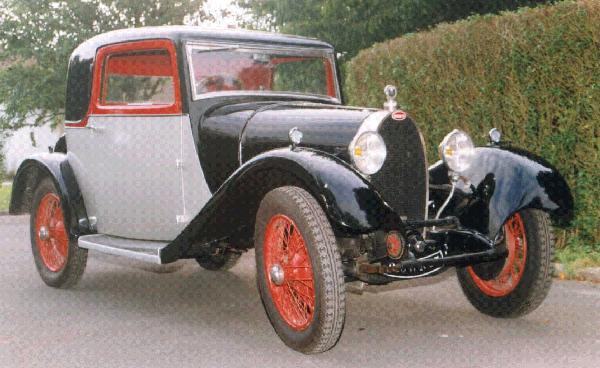 Pistonheads - The image shows an antique car with a classic design. The car is primarily gray with white trim, and it features a black roof. It has distinctive red and orange wheel rims, which contrast with the rest of the vehicle. The car is parked on a street next to a green hedge, indicating it's in an outdoor setting.