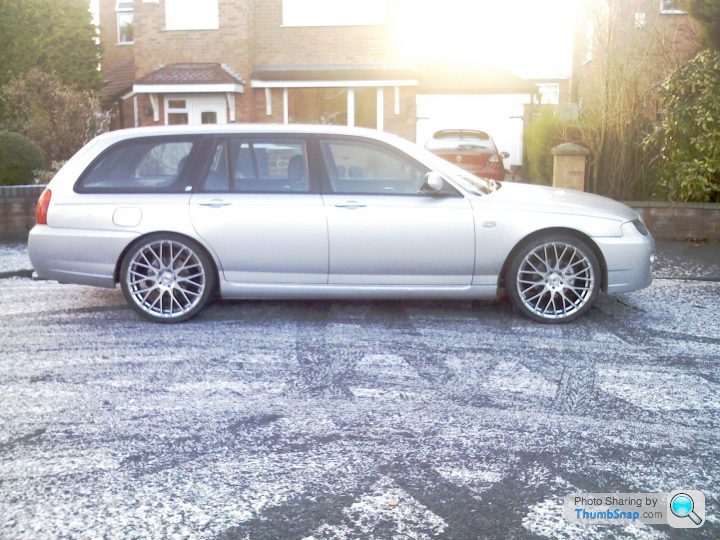 Service Costs Pistonheads - The image shows a silver-colored station wagon stationed on a driveway of a residential area. The car has a shiny finish and large, chrome rims. A car is parked behind the station wagon, and a garage door can be seen in the background. The time of day appears to be near sunset, as there is a warm glow from the sky.