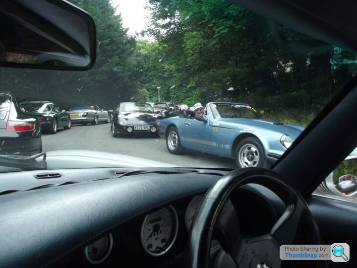 TVR,s at Chatsworth. Saturday runs October 3rd - Page 1 - TVR Events & Meetings - PistonHeads - The image captures a scene from inside a black car, where traffic is moving slowly along a tree-lined road. The road is filled with a variety of vehicles, including a teal convertible and several black cars. The view through the car window also shows the edge of another vehicle, suggesting the perspective of someone in the back seat of the black car. There are several trees, providing a green canopy over the road, and the overall atmosphere appears to be bright, possibly suggesting it's daytime.