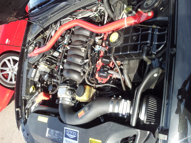 *Forum Event* Performance Vauxhall Show 2012 - Page 9 - HSV & Monaro - PistonHeads - The image shows a hood of a car, which is open, revealing a complex assortment of engine components. The engine is composed of various parts including a red plastic part, a black air filter, and a metal exhaust pipe. There's also a red engine hose near the exhaust pipe. On the trunk of the car, there's a GTO emblem. The engine bay appears to be well maintained, indicating that the car is likely in good condition.