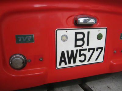 Early TVR Pictures - Page 35 - Classics - PistonHeads - The image captures a Red Volkswagen bus parked on a street. The license plate of the bus prominently displays the letter "B" and the numbers "I AW577". The license plate is secured by a chrome plate with a locking mechanism on either side of the plate. The bus appears to be parked at the curb, and the background provides a glimpse of the street. There is a hint of another vehicle in the far distance. The license plate is the central focus of the image, providing detailed information about the vehicle.