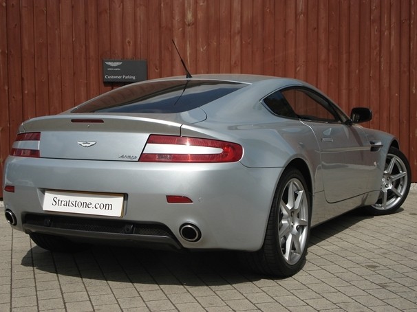 Pistonheads Guys Owner - The image features a silver Aston Martin car parked on a brick driveway. The vehicle is oriented facing away from the camera, showcasing its sleek rear design. The Aston Martin logo is prominently displayed on the trunk, confirming the brand of the car. Above the car, a vertical sign indicates "CUSTOMER PARKING", suggesting that this is a designated parking spot for customers. The background includes a wooden wall, adding a rustic charm to the overall scene.