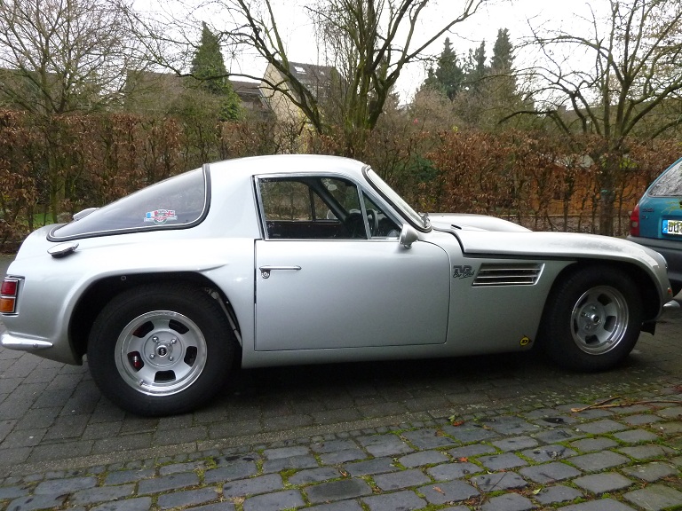 Early TVR Pictures - Page 92 - Classics - PistonHeads - The image depicts a silver, open-top sports car parked in an outdoor space, which appears to be a driveway. The vehicle is a two-seater with a long nose, a design that is characteristic of classic sports cars. A black credit card style decal on the car's rear window prominently features a flag with a red cross and other decorative elements. The surrounding environment includes a variety of leafless trees and some bushes, indicating it might be autumn or winter. Another vehicle is parked in the background, and the ground is covered with small cobblestones.