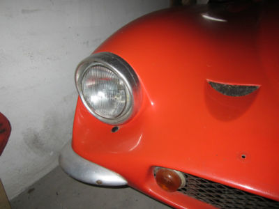 Early TVR Pictures - Page 35 - Classics - PistonHeads - The image displays the lower part of an orange car with a prominent front bumper, faux grille, and a headlight. The car is parked, with its body taking up most of the frame. The background is simplistic, featuring a concrete or cement area suggesting an outdoor parking space. There are visible dust or smudges on the base of the car, and a small part of the hood can be seen at the bottom. The style of the image is candid and utilitarian, possibly taken during a search or inventory process, focusing primarily on the details of the car's front end.