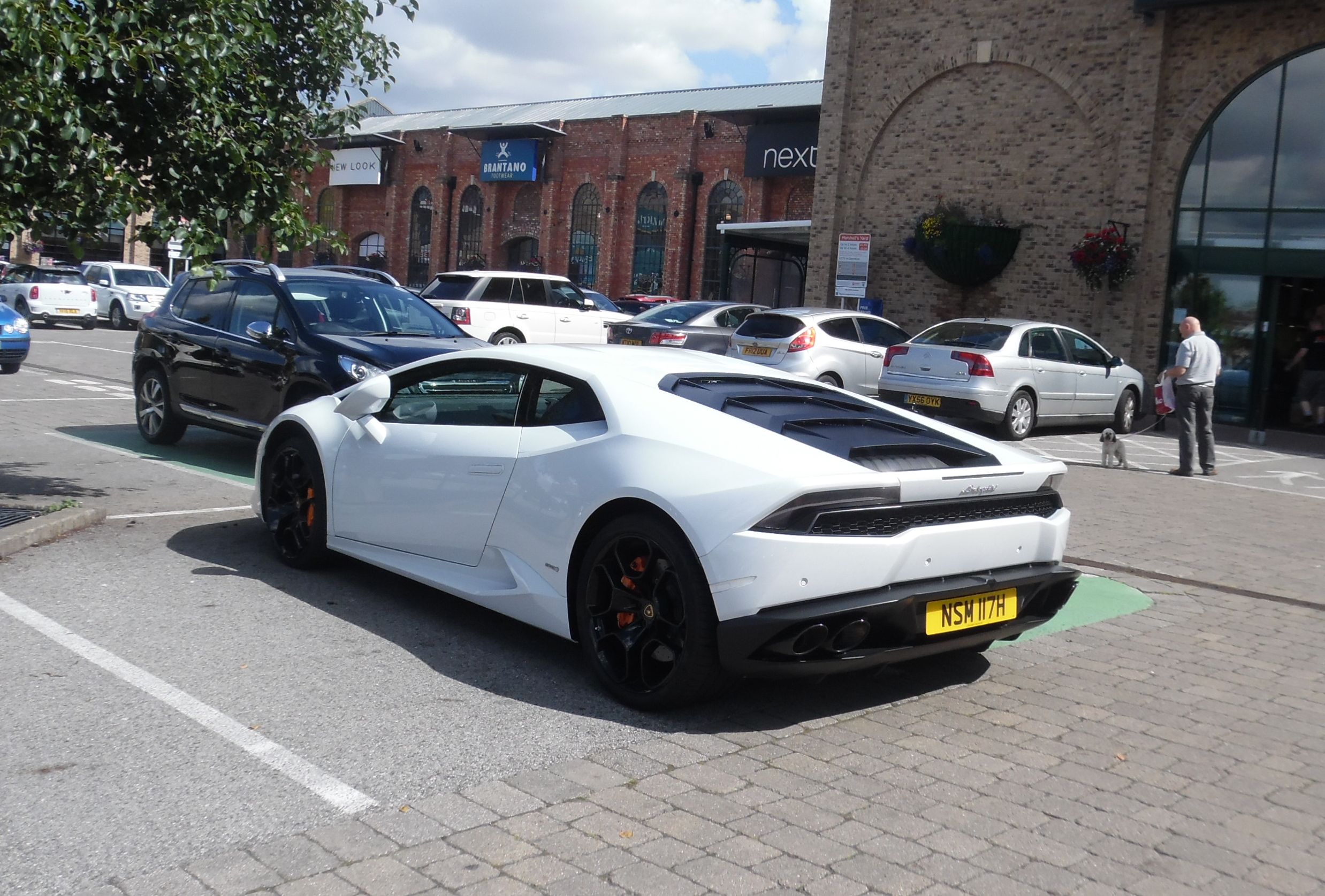 Midlands Exciting Cars Spotted - Page 324 - Midlands - PistonHeads - The image shows a white sports car parked in a parking lot, which appears to be located in a shopping area given the presence of a building with storefronts in the background. The car is angled slightly to the side of the frame and takes up a large part of the image. It has distinctive design elements, such as large aerodynamic vents on the side and a pronounced rear wing. The vehicle appears to be modified for performance with visible aftermarket parts. There are also other cars visible in the background, but the sports car is the main focal point of the image.