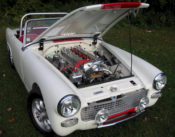 Pistonheads Swap Ideas Engine Mgb - The image shows a vintage white convertible car with the hood up, revealing an engine with visible red connectors and various metallic components. The setting appears to be a grassy area with a natural backdrop, indicating an outdoor photo shoot. The car's design features traditional early-to-mid 20th-century styling, with round headlights and a distinct front grill. The hood is held propped up, offering a clear view of the complex mechanical components typical of vehicles from that era. There are no visible texts or logos in the image.