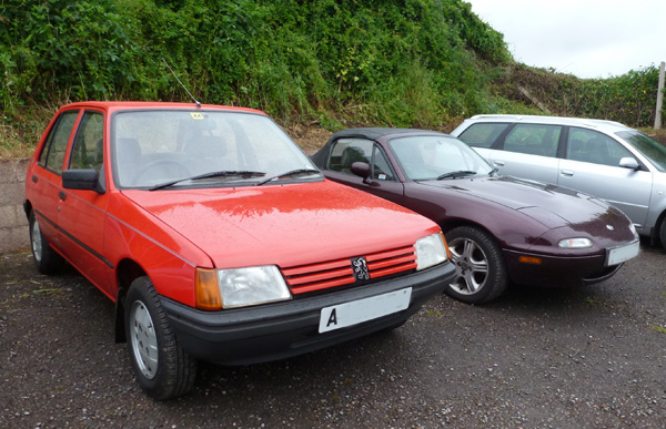 Local meet - Page 1 - South West - PistonHeads - The image shows a parking lot scene with three cars parked on an asphalt surface. In the foreground, a red Volkswagen Beetle is prominent, covered in some spots with car wax or a similar reflective substance. To the right stands a black two-door sports car with a distinct shape and design, and behind it is a silver sedan, larger in size and more conservative in appearance. The backend of an additional car is visible in the upper right corner, but it's obscured and not clearly identifiable. The setting appears to be a paved lot amidst lush greenery, suggesting a suburban or semi-rural environment. There's a gate or fencing area in the background behind the silver car. The sunlight seems to be uneven, casting shadows between the cars, indicating an outdoor setting with natural light.