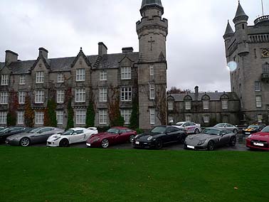 Pistonheads - The image shows a group of cars parked in front of a stately, castle-like building with towers and battlements. The cars vary in color and model, and they are arranged along the curb of a lawn. The weather appears overcast, with a pale blue sky suggesting either dawn or dusk. The setting is reminiscent of a moveable period drama with the cars possibly representing different time periods. The building's architecture is grand and ornate, very unlike the modern design of the cars.