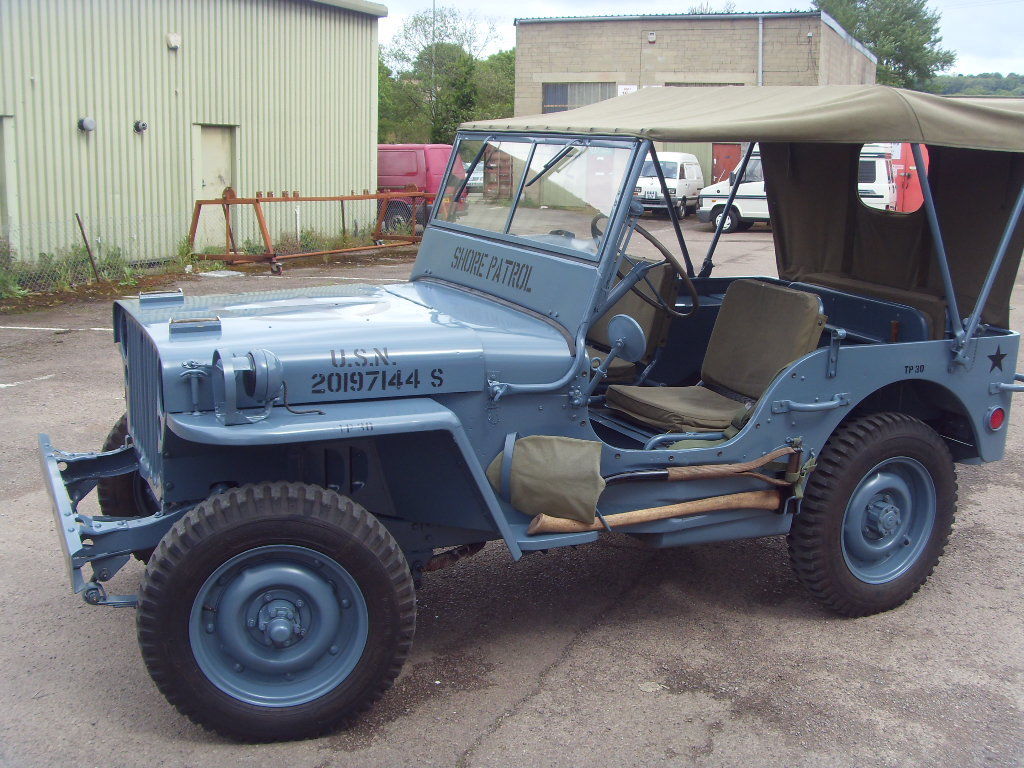 New Family Member: 1943 Ford GPW Jeep! - Page 1 - Classic Cars and Yesterday's Heroes - PistonHeads - In the image, there's a unique sight of an older, military-grade jeep stationed in a gravel lot. The vehicle is painted in a color that's a blend of grey and blue. It's positioned parallel to the camera, giving a full view of its front and side. The jeep has an eight-sided hood, a feature characteristic of World War II jeeps, and its body appears to be constructed from wood. Several small windows are visible on the sides, which suggests that the vehicle was designed for use in various weather conditions and to accommodate passengers or workers. The blocky shape of the jeep is reminiscent of the military vehicles of an earlier era.