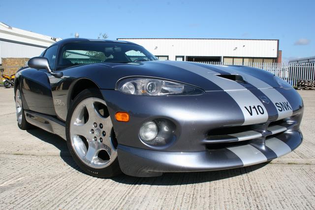 Newbie  - Page 1 - Vipers - PistonHeads - This image shows a sports car parked on a paved surface, likely in a commercial or industrial setting given the presence of a building and a fence in the background. The car is a sleek, two-door model with a modern design, featuring a black and silver color scheme on the hood, and is labeled with "V10" and "SKW." The vehicle is positioned such that the hood appears slightly raised, and the lighting suggests it may be parked in a sunnier location, with the car's metal body reflecting some of the sunlight.