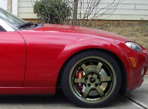 which alloys for my MK3 - Page 1 - Mazda MX5/Eunos/Miata - PistonHeads - The image features a close-up of the front wheel of a red, convertible sports car. The car has a polished wheel that is black in the wheel wells and brown in the centers. The polished edge of the fender extends over the top of the tire, a characteristic design element that gives the vehicle a sleek and sporty appearance. The environment around the car includes a pavement surface and a grassy area with a tree in the background. The sky is overcast, suggesting a cloudy day.