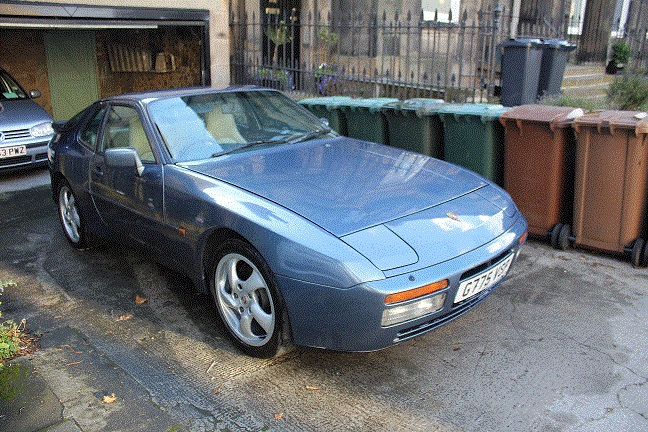 Just picked up my daily runner, she's a looker!! - Page 1 - Front Engined Porsches - PistonHeads - The image depicts a vibrant blue sports car parked in front of a building. The car's glossy finish reflects the sunlight, and it is positioned on top of a gray driveway. The setting is urban, with a stack of trash cans visible in the background to the right. Adjacent to the trash cans, a green recycling bin is placed next to a building fence. Further in the distance, another vehicle is visible on the left side of the frame. The car gives off a sense of luxury and sportiness, contrasting with the mundane domestic background.