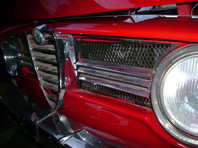 Bertones Beautiful Pistonheads - This image captures a close-up view of the headlight of a red classic car. The headlight is illuminated and its glass facet is visible, revealing the intricate details of the light. The car appears to be a vintage model, as suggested by the classic design and the sturdy grill enclosing the headlight. The focus of the image is on the headlight itself, highlighting its condition and the depth of the car's color. The vibrancy of red on the car's hood adds a striking contrast to the metallic details of the vehicle.