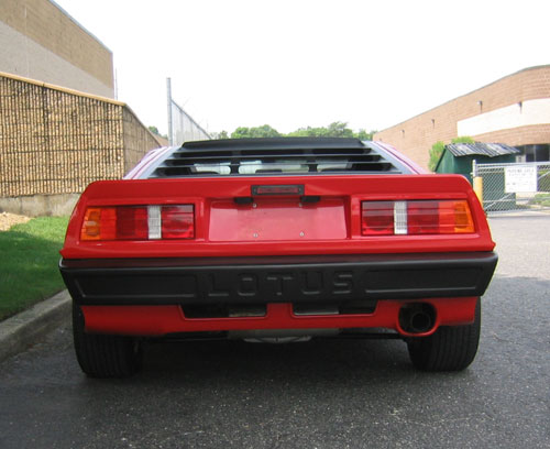 Pistonheads Shared Components - The image shows a classic red Lotus sports car parked on a paved surface next to a green fence. The rear of the car is visible, featuring a distinctive aerodynamic body design with a small black spoiler on the rear deck. The vehicle has horizontal red and white brake lights, with the brand name "Lotus" prominently displayed in the center rear panel. The backdrop is relatively nondescript, focusing on the striking color and unique shape of the Lotus sports car.