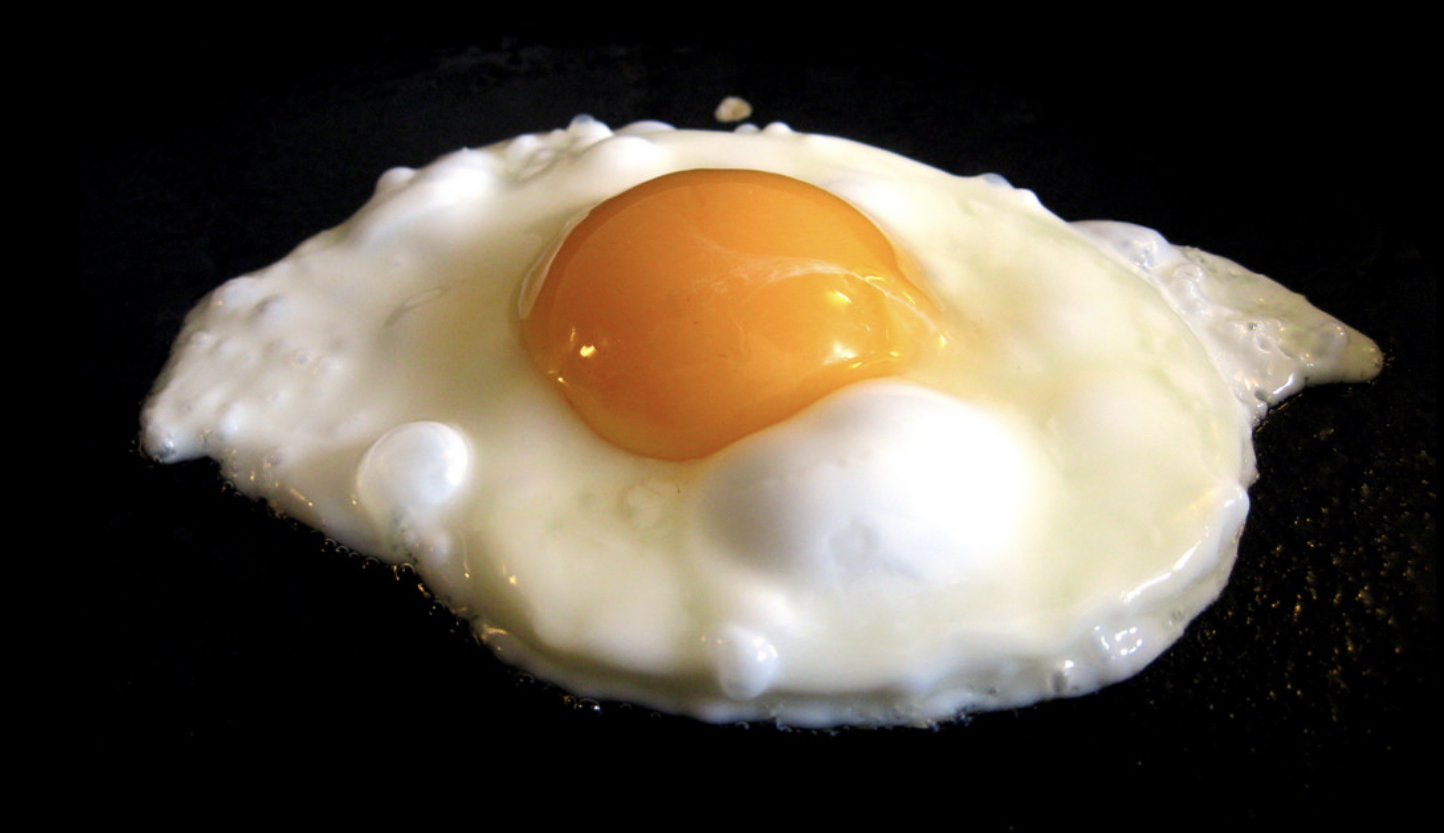 Nicknames for the best version - Page 5 - Classic Cars and Yesterday's Heroes - PistonHeads - The image shows a single, freshly-made fried egg sitting on a dark surface. There's a yellow yolk visible in the center of the egg. In the background, there is a blurred depiction of an electric stove or hot plate with a pan. To the right of the egg, there's a small amount of water and a drop of oil, which were likely used in cooking the egg.