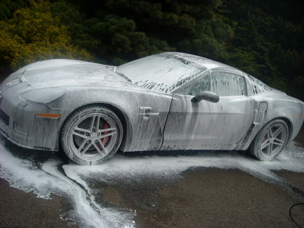 Properly Clean Pistonheads - The image features a dark-colored sports car covered in a thick, white foam which appears to have been applied by a pressure washer. The car is parked on a dark surface, possibly a tarmac or blacktop, and the foamy substance is dispersed attractively on the ground, creating a stream and puddle effect around the vehicle. The car's glossy finish is partly obscured by the foam, and the stream from the pressure washer suggests that the car is being cleaned or transformed in some way.