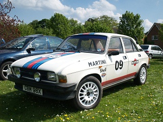 Foxhall Thunder 500 Pistonhead Roadshow - Page 1 - East Anglia - PistonHeads - The image features a striking Martini car parked on a grassy field. The car is predominantly white, with a red and blue stripe running down the side, a theme commonly associated with Martini. The vehicle displays the Martini logo and the number 09, suggesting it may be part of a collection or a racing team. In the background, other cars can be seen, possibly indicating a larger gathering or event. The setting appears serene and outdoor, with trees visible in the distance, adding a natural element to the scene. The weather appears clear, suggesting good conditions for a potential car-related event.