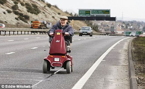 If its Green It Needs to be Seen.. - Page 1 - Aston Martin - PistonHeads - In the image, an elderly gentleman is seen riding a small, red scooter on a multi-lane road during the day. The person is wearing a cap and glasses, suggesting mild weather conditions and a need for proper vision. The road is relatively clear with only a few other vehicles, mainly cars and trucks, present. The overall scene appears to be a typical day on a highway, with the scooter rider as the main focus.