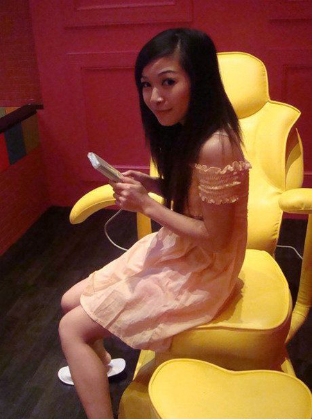 The image showcases a woman in a vibrant room with yellow and red accents. She is seated comfortably in a plush yellow velvet chair, holding a remote control in her hands, suggesting she might be in a gaming or entertainment setup. Her dress is pink, blending well with the warm-toned room. She appears to be in a relaxed, casual setting, possibly in her own home or retreat.