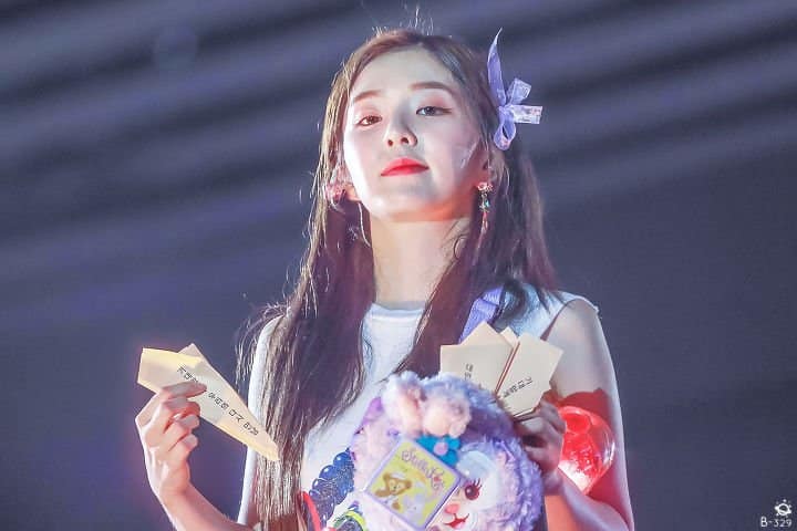 A woman holding a cell phone in her hand - In the image, a woman stands out against a dark background. She is elegantly dressed in a white top and is adorned with a blue headpiece. In her hands, she holds two thoughtfully crafted items: a purple clutch resembling a flower and a white book with text. Her pose and the items she holds suggest she might be presenting these items, possibly in a musical performance given the look on her face.