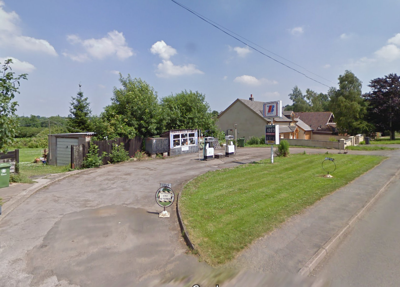 The Humer Unbeam Interesting Filling Stations Thread - Page 46 - General Gassing - PistonHeads - The image shows a tranquil street corner during the day. On the right side of the road, there are houses in the background, covered with green foliage. The left side of the road has a street sign visible. There is a small black and white sign with a directional arrow pointing left, standing in grass next to the road. The pavement is smooth and well maintained. The sky above is clear with a few clouds scattered across it.