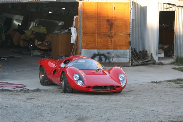 tempero cars - Page 1 - New Zealand - PistonHeads - The image shows a striking red vintage sports car parked on a dirt lot next to a building with orange metal doors. The car features a prominent air intake on the front, along with prominent headlights and a curved windscreen. A white "X" is drawn on the orange metal door of the building. There are metal pieces in the background, suggesting an industrial or workshop setting. The perspective of the image is from a low angle, looking up at the vehicle, emphasizing its sporty design.