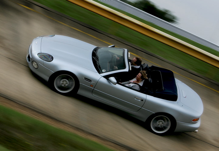 Your DB7 pictures. - Page 1 - Aston Martin - PistonHeads - This image captures an exhilarating moment of a silver convertible car in motion. The car is driving on a highway with a blurred background, suggesting speed. There are two individuals enjoying the ride, their hair splayed by the wind due to the open top of the car. The angle of the image gives a dynamic impression of the car's movement down the road.