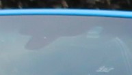 Liber T Tag and possible windscreen Issue? - Page 1 - Boxster/Cayman - PistonHeads - The image shows a close-up of a blue vehicle's windshield. Through the glass, the silhouette of a person dressed in dark clothing is visible. The angle and composition create a sense of mystery, as the person's identity is obscured by the reflection in the windshield. The focus is sharp in the foreground, capturing the clear blue of the windshield and its reflection of the sky. The image has a dramatic nature, with the contrasting the vibrant color of the windshield against the dark background of the sky.