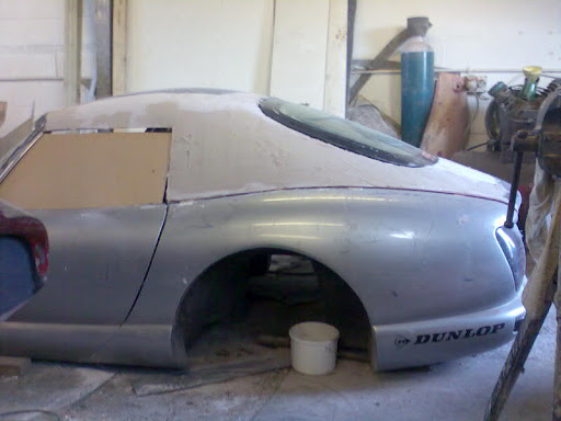 Cerbera Style Hard Top for Chimi´s - Page 1 - Chimaera - PistonHeads - The image shows a silver-colored car that is in the midst of a renovation or repair process. The car is taped off in the garage, indicating that it's being worked on. It is positioned on a hydraulic lift, suggesting that the undercarriage or rear end is being worked on. A red bucket is visible near the car, possibly containing tools or materials for the repair. The background appears to be a garage or workshop setting with additional tools and equipment visible.