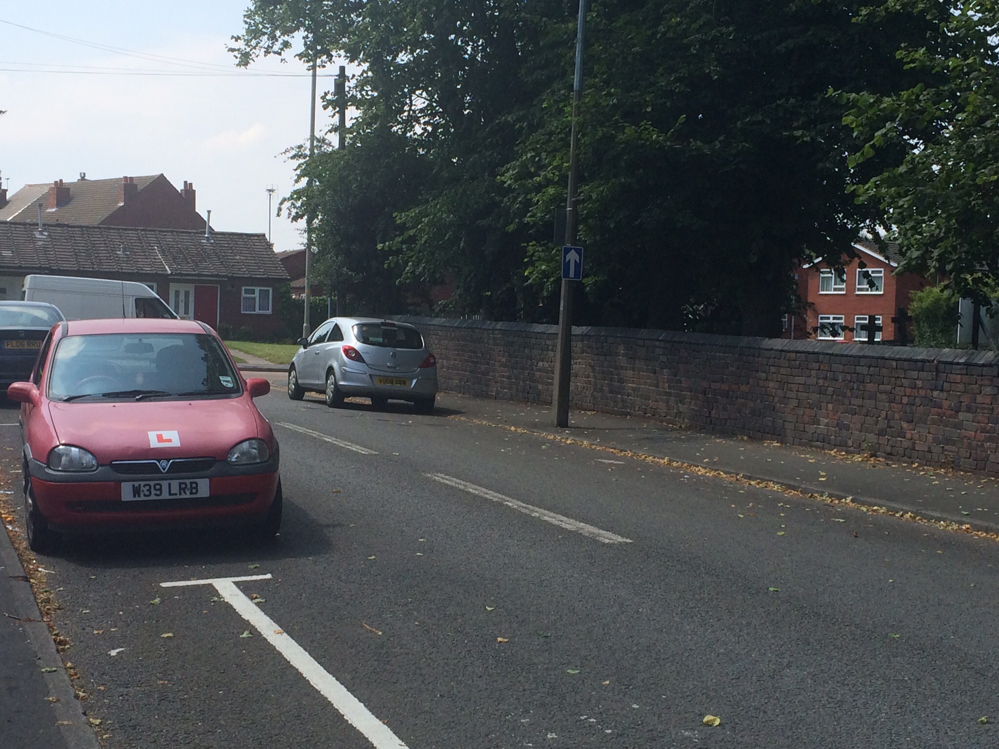 The BAD PARKING thread [vol3] - Page 27 - General Gassing - PistonHeads - The image depicts a quiet residential street lined with brick walls and mature trees. Parked along the pavement are two cars - a vibrant red hatchback with a white license plate and a metallic grey car, both facing to the right, possibly parked on either side of a driveway leading to a red-roofed house. The scene conveys a sense of tranquility, typical of suburban areas.