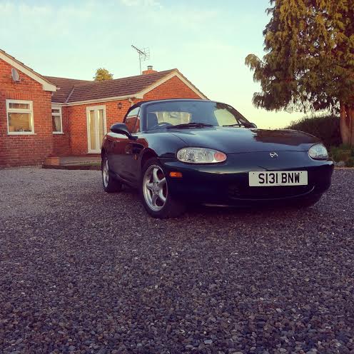 My new MX-5!  - Page 1 - Mazda MX5/Eunos/Miata - PistonHeads - The image shows a black Mazda Miata parked in front of a house. The car's model and license plate are clearly visible. It's a sunny day with a partly cloudy sky. The ground features a gravel driveway. The house in the background has a brown exterior.