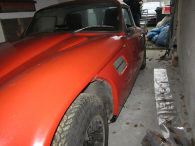 Early TVR Pictures - Page 35 - Classics - PistonHeads - The image depicts a dark red or orange vintage convertible car with a prominent front fender and a sleek, shiny surface. The vehicle appears to be parked inside a garage or similar indoor space, as it is situated on a cement floor with other miscellaneous items visible in the background. The car's design suggests it's a 4-door model but the hood doesn't seem to be fully visible or in place. There are no texts or specific information discernable from the image itself. The style of the car indicates it may be from the 1960s or earlier based on its body lines and overall aesthetic.