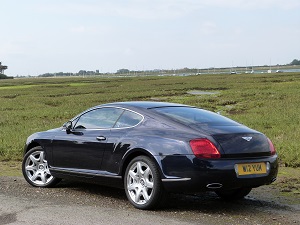 New purchase and thanks - Page 1 - Rolls Royce & Bentley - PistonHeads - In the image, a sleek black sports car with a yellow license plate is prominently parked on the side of a road. The vehicle is shiny and well-maintained, indicating it's in good condition. The road it's parked on appears to be made of dirt or gravel, suggesting a rural or less-traveled area. Beyond the car, there's a picturesque landscape filled with green fields and at a distance, a line of trees or bushes can be seen, adding to the serene and natural beauty of the scene. The car's design and the surrounding environment suggest a sense of luxury and openness.