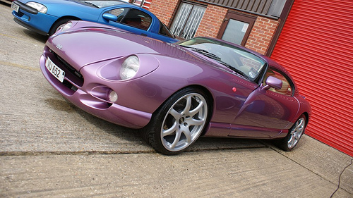 Our Cars - Page 39 - South Coast - PistonHeads - The image shows a scene with two vintage sports cars parked next to each other. The car in the foreground is a purple sports car with a sleek design and chrome detailing, positioned on a road partially covering the curb. To the right of this car, there is a bright blue sports car, partially visible behind it. In the backdrop, immediately behind the cars, there is an old brick building with a red shutter. The ground appears to be constructed of concrete or asphalt, and there is a sense of age and history associated with the vehicles and the building. The overall atmosphere of the image suggests a classic, nostalgic theme likely meant to celebrate or evoke the spirit of vintage automobiles.