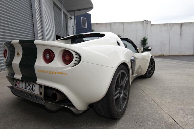 Elise Pistonheads Lotus - The image shows a sleek, white sports car parked on a concrete surface. The car features a distinct black stripe running along its side, and two red brake lights on the back. The vehicle has a low profile with stretched bodywork, consistent with tuning modifications. The setting appears to be a business area, as suggested by a fence and a building with roll-up doors in the background. The sky is partly cloudy, indicating a change in weather.