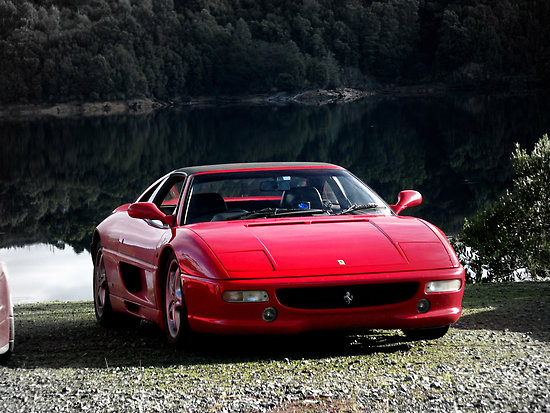 Is this fake? - Page 1 - General Gassing - PistonHeads - The image features a sleek red Ferrari sports car parked on a gravel road or bank. The car is positioned diagonally facing the left side of the photo, with the tranquil and reflective body of water adjacent to it. The surrounding landscape includes dense green foliage, and uphill in the background, the silhouette of a mountain range can be seen. The overall impression is one of luxury, in a natural and serene setting.