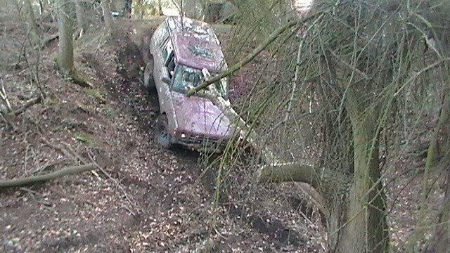 Wilton off road centre pics 11/03/2012 - Page 1 - Off Road - PistonHeads - The image depicts a purple vehicle that has veered off a roadway into a wooded, steep area with tall brush. The vehicle appears to be old and dirty, suggesting it may have been abandoned or accidentally brought into this environment. The surrounding scenery is dense with branches and tree stumps, adding to the sense of isolation and abandonment. There are no people visible in the immediate vicinity of the vehicle. The forest setting indicates a remote location, which might be in the countryside or a less traveled area. The photo seems to have been taken with a camera equipped with a zoom feature due to the close-up angle and the red and pink hue of the image, which could be the result of a filter or lens.