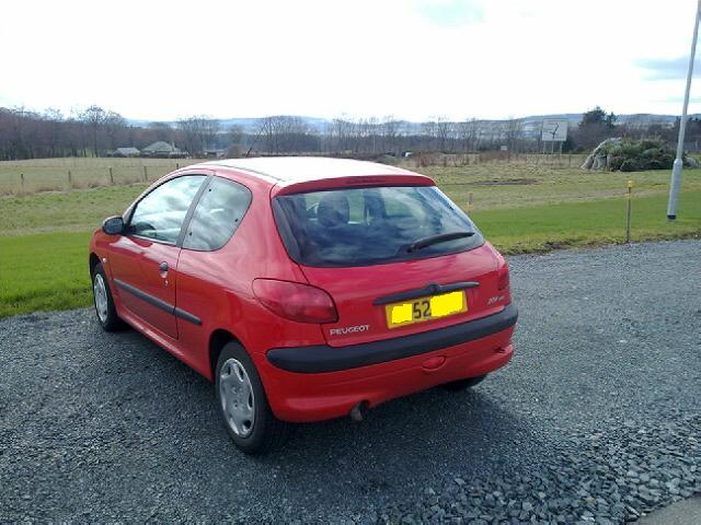 Up yours Gordon ! - Page 1 - Readers' Cars - PistonHeads - This image features a red hatchback parked on a gravel area near a field. The vehicle has a license plate visible on the back and appears to be a standard model with side mirrors, a visible antenna, and a design that suggests it's a late-model year. The sky is overcast, suggesting this photo was taken on an overcast day. The field in the background appears lush and well-maintained, which could indicate a rural or suburban setting.