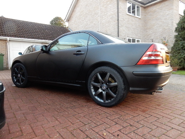 Progress thread 2001 Mercedes SLK230 - Page 3 - Mercedes - PistonHeads - The image displays the rear view of a dark-colored luxury sedan parked on a brick driveway. The vehicle has tinted windows and five-spoke custom-painted alloy wheels, giving it a sleek and sporty appearance. The sedan is one of the newer models, identifiable by the chrome emblem on the tail light and the license plate above the muffler. The side mirrors and detailing give the car a polished and personalized look.