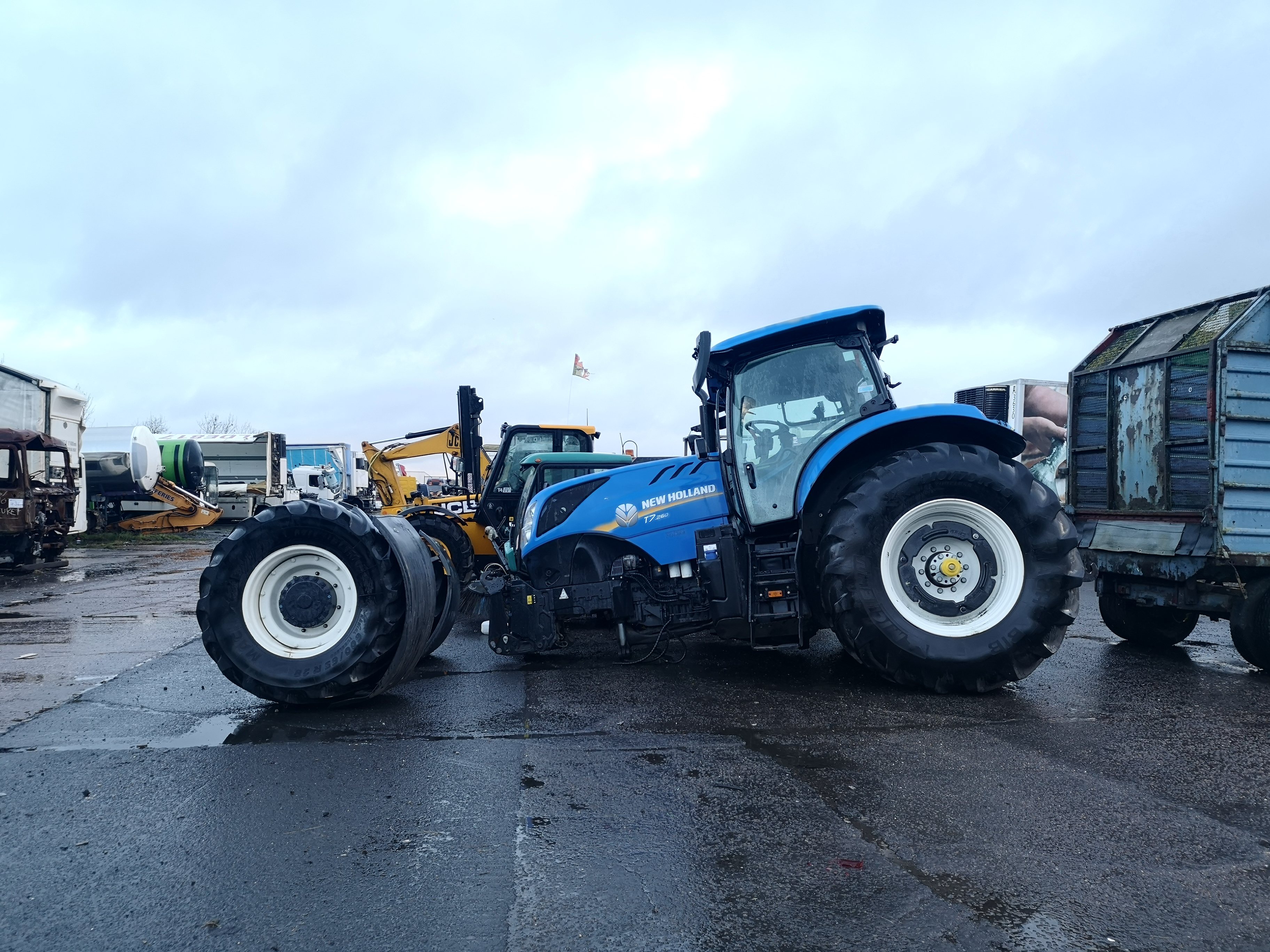 Show us your crash pics!! - Page 347 - General Gassing - PistonHeads UK - The image showcases a scene in what appears to be an industrial or commercial setting. A blue tractor is parked prominently in the foreground, its large wheels and sturdy build indicative of agricultural machinery. Behind the tractor, other vehicles are visible, including a truck with a flat bed. These vehicles suggest activity related to transportation or construction. The ground is wet, hinting at recent rain, which has left puddles on the asphalt surface. Despite the industrial setting, there's a touch of life represented by the presence of several birds scattered across the image, adding a dynamic element to the scene.