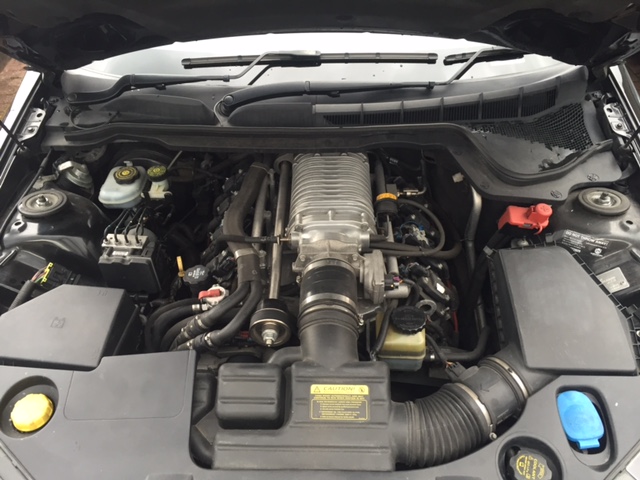 Older charger not intercooled - Page 1 - HSV & Monaro - PistonHeads - This image shows the engine bay of a vehicle, likely an SUV given the size of the engine and the black color scheme. The engine appears to be a V8, as indicated by the number of cylinders and the overall layout. The motorbelt is made of black rubber and runs across the top of the engine. There are various components, such as hoses, filters, and other mechanical parts, which are typical in an engine bay. The background is darkened, possibly due to the car's interior or the angle of the photograph.